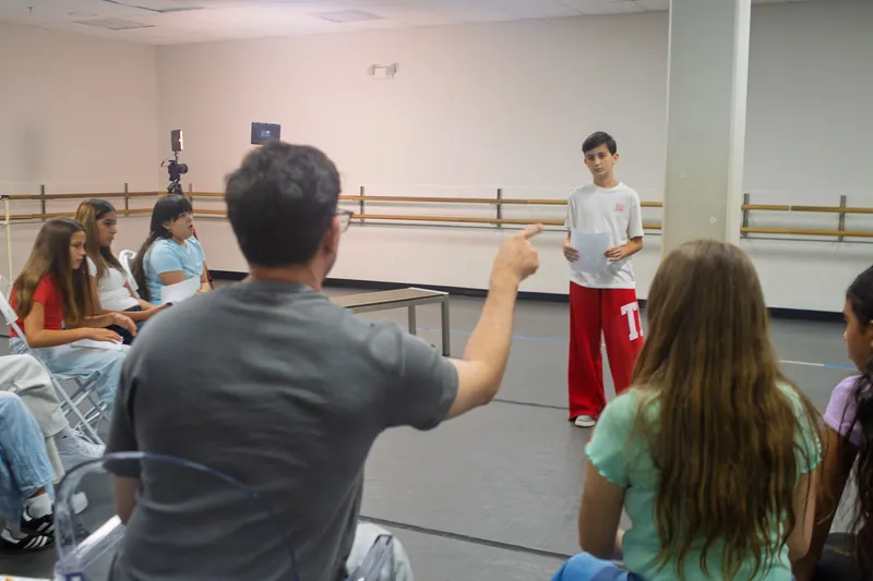 Seen from behind, Paul Becker pointing across the studio to a student performing a scene while classmates watch from chairs